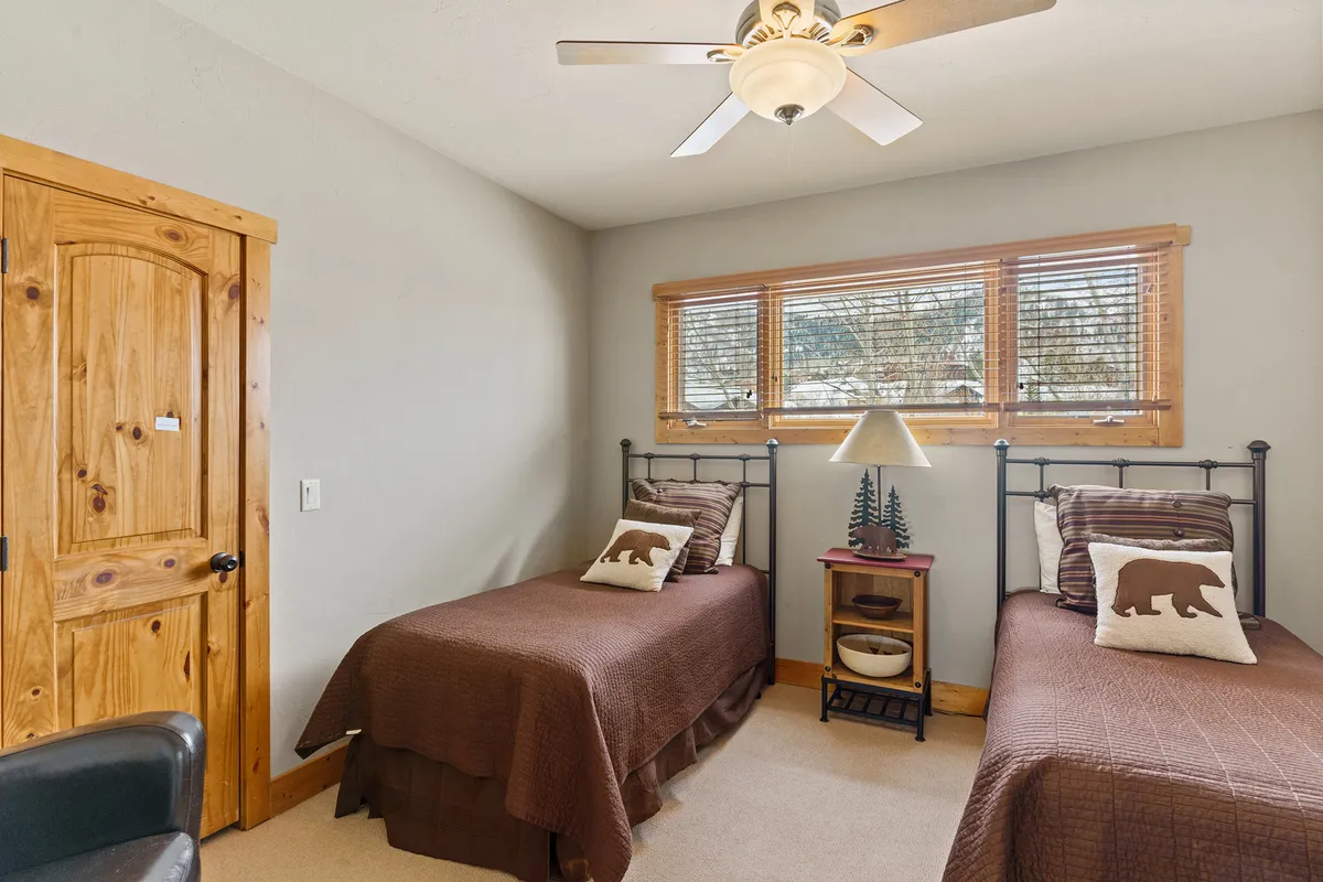 A bright and comfortable bedroom with twin beds, a wooden door, window with blinds, and a nightstand with a lamp. The setup suggests a welcoming space ideal for children or friends, offering both comfort and a sense of togetherness.
