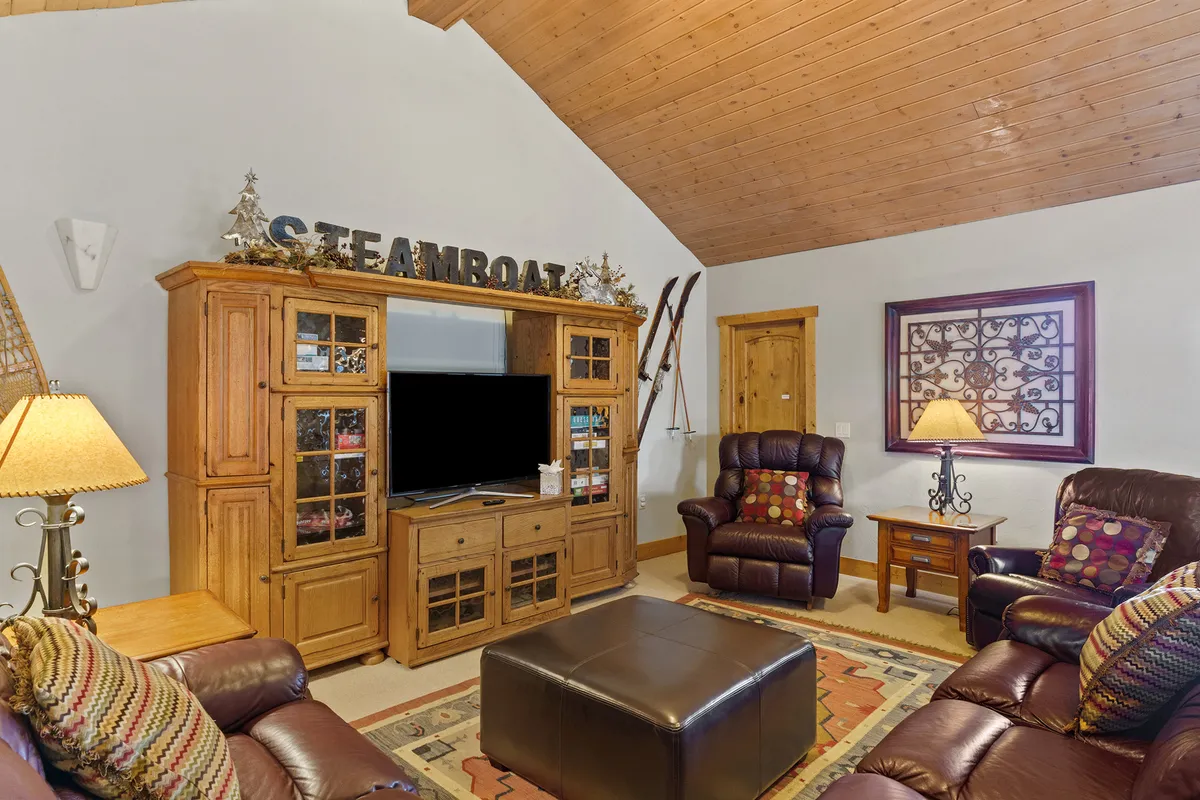 A cozy lounge area anchored by a wooden entertainment center, reclining chairs, a coffee table, and soft lamp lighting. Decorative wall art and comfortable seating provide a relaxed atmosphere ideal for watching TV, playing games, or enjoying snacks from the wet bar.