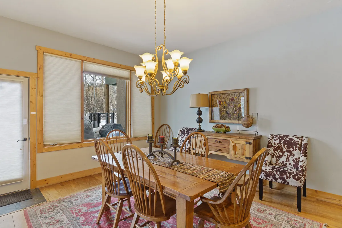 This image features a welcoming dining area with a central table, a statement chandelier, decorative chairs, and a window that offers a bright outside view. The sideboard with decor adds a homely, festive touch, suggesting an inviting space for gatherings.