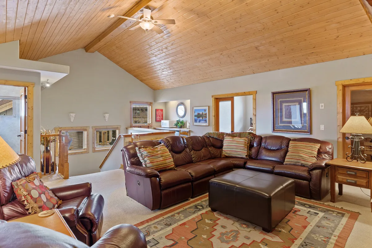 A spacious upstairs family room featuring a large sectional sofa with decorative pillows, a coffee table in the center, a warm wooden ceiling, and framed artwork on the walls. The setup invites group lounging, movie nights, and casual gatherings, encapsulating the heart of a family fun zone.