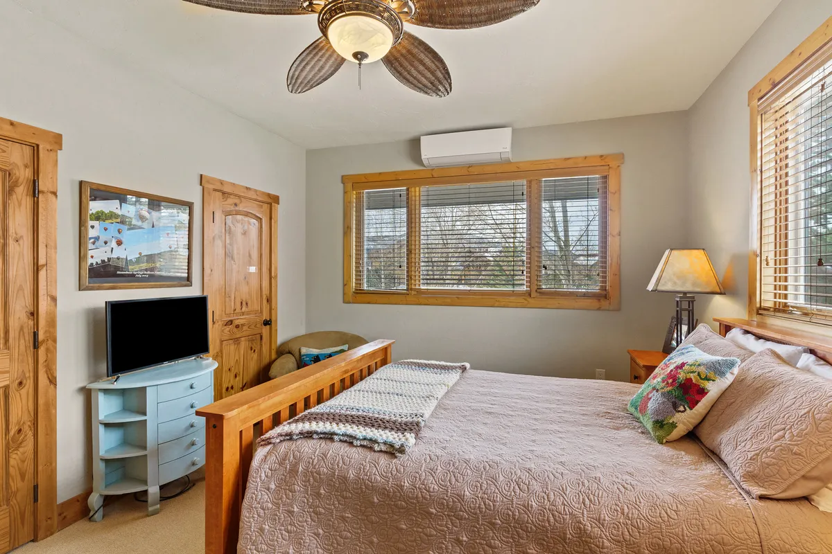 A serene bedroom featuring a bed with decorative pillows, wooden furniture, and a window with blinds. A TV is positioned on a stand near the bed, and a lamp beside the bed offers cozy illumination. The room feels spacious, comfortable, and equipped with modern amenities.