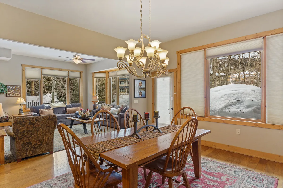 A cozy dining space featuring a wooden dining table under an elegant chandelier, set beside large windows that let in abundant natural light. The comfortable seating area nearby creates an inviting spot for family dinners or casual brunches.
