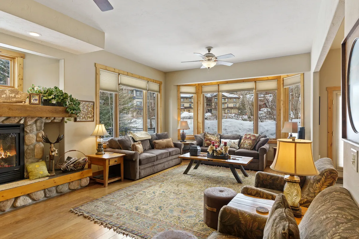 This cozy living room features a stunning stone fireplace, expansive windows letting in mountain light, and plush seating arranged around a coffee table decorated for comfort and style. The space exudes warmth and invites families or groups to gather, relax, and enjoy each other's company in a mountain retreat setting.